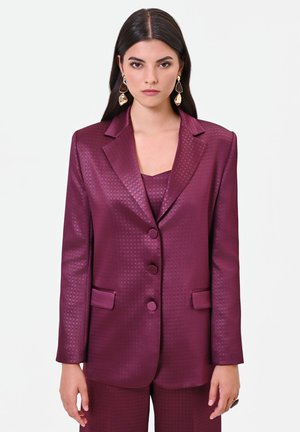 Femme aux cheveux lisses et foncés porte un blazer texturé brillant couleur prune et un pantalon assorti avec des boucles d'oreilles dorées remarquables.