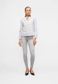 Νεαρή γυναίκα στέκεται φορώντας λευκό μπλουζάκι με λουλούδια, ανοιχτογκρι σκισμένα στενά τζιν και μαύρες μυτερές γόβες μπροστά σε λευκό φόντο.