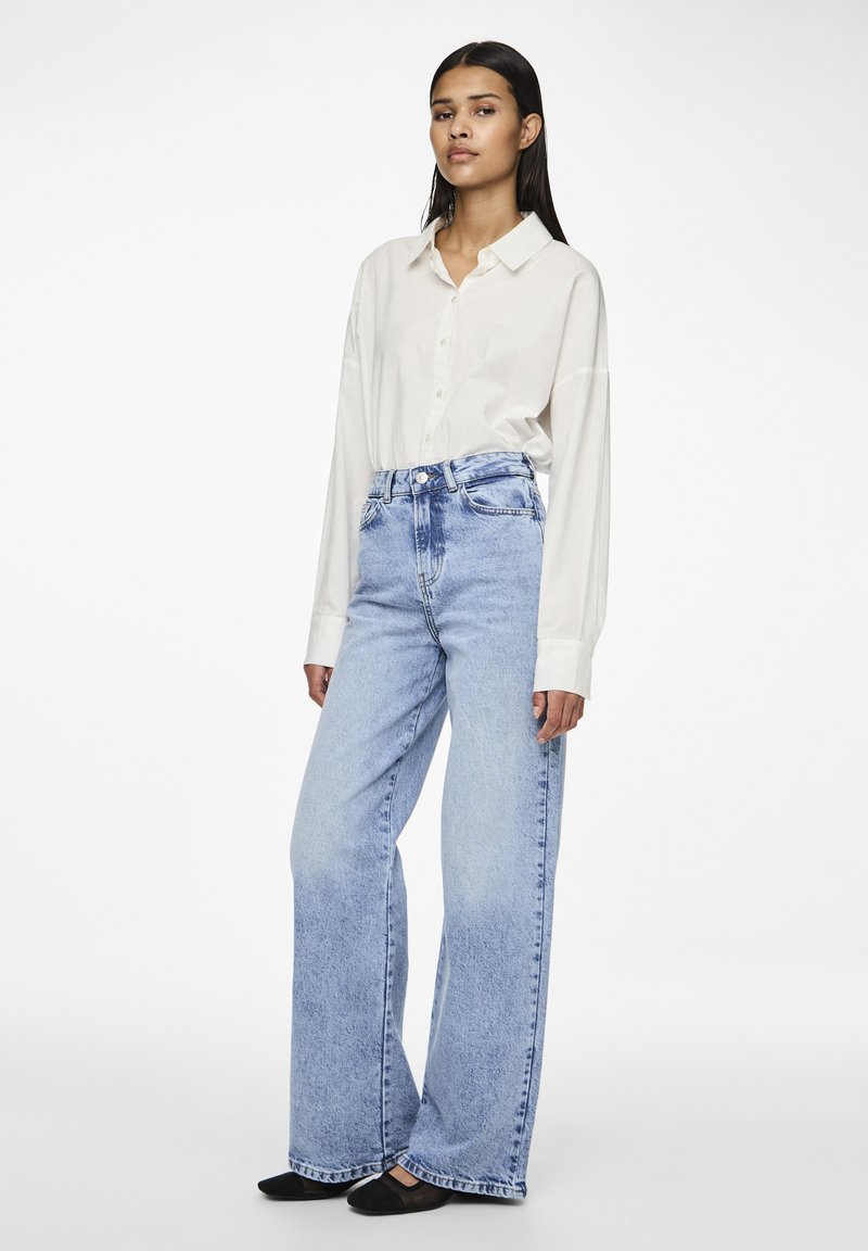 Losse lichtblauwe spijkerbroek met hoge taille, gecombineerd met een off-white blouse met lange mouwen en zwarte platte schoenen.