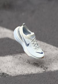 Lightweight, off-white running shoe with textured upper, blue accents, and a cushioned sole. Features laces and a prominent Nike logo.