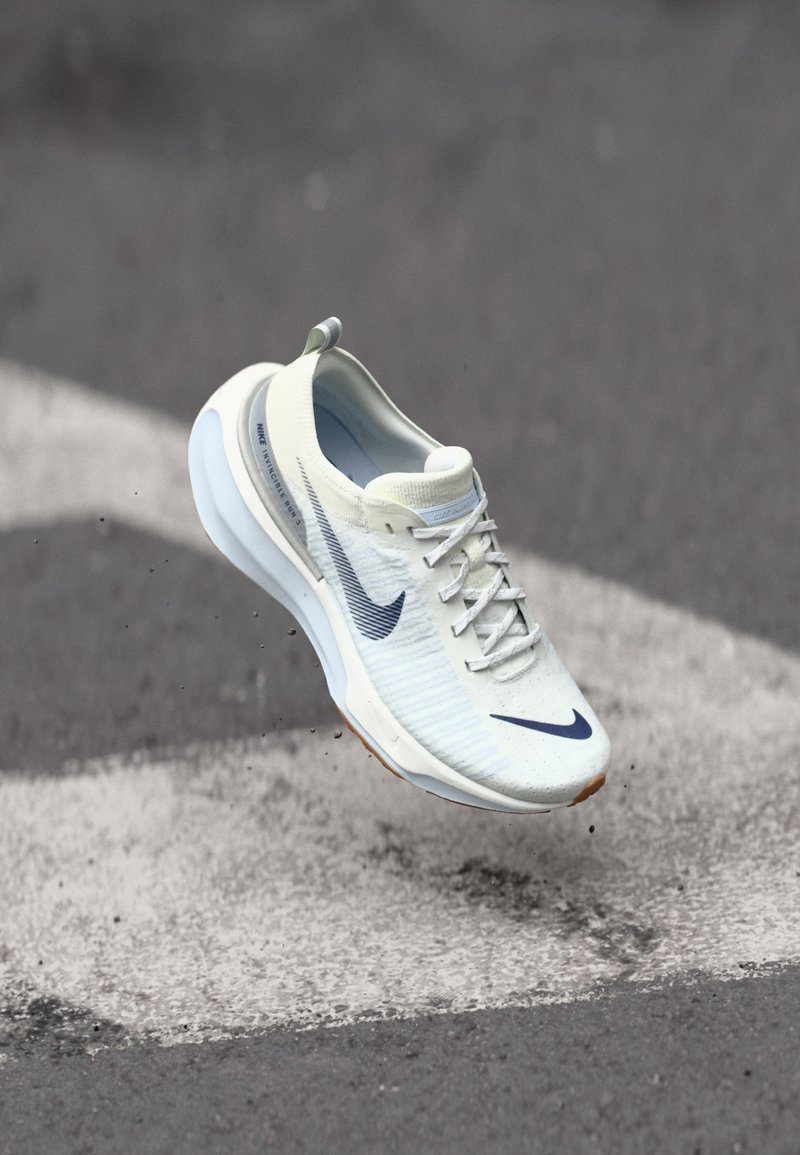 Lightweight, off-white running shoe with textured upper, blue accents, and a cushioned sole. Features laces and a prominent Nike logo.
