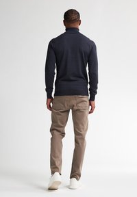 Maglione a collo alto navy con maniche lunghe, abbinato a pantaloni khaki. Il modello indossa scarpe da ginnastica bianche, in piedi contro uno sfondo neutro.