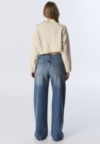 Femme aux longs cheveux portant un pull court en tricot torsadé crème et un jean en denim bleu à jambes larges, se tenant devant un fond léger uni.