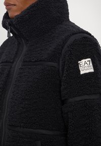 Svart fleecejacka med hög krage, texturerat tyg, kontrasterande släta paneler och en EA7 Emporio Armani-logotyp på ärmen.
