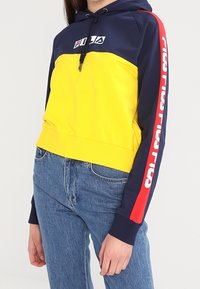 Felpa con cappuccio a blocchi di colore con corpo giallo e cappuccio blu navy, con un accento rosso sulla manica e logo bianco. Materiale in misto cotone, design corto.