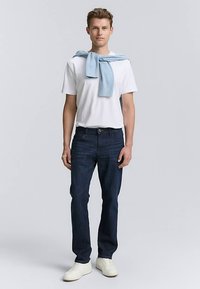 Balti marškinėliai, tamsiai mėlynos džinsai, šviesiai mėlynas megztinis, surištas ant pečių, ir balti sportbačiai. Stovi pozicija priešais vienspalvį foną.