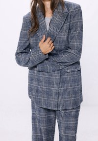 Blazer a cuadros en tonos azul marino y gris, con un diseño estructurado, solapas anchas y cierre frontal superpuesto con un corte holgado.