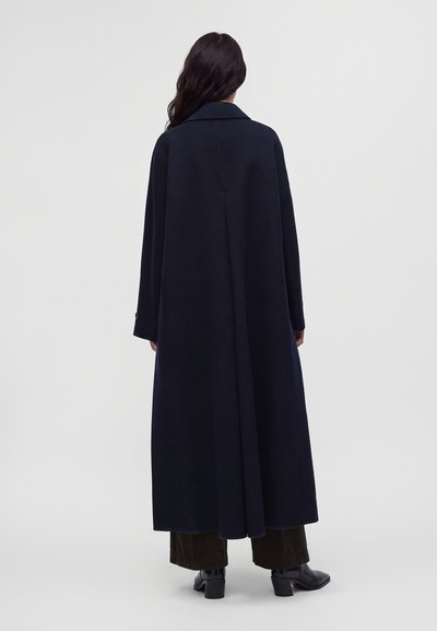 Femme aux longs cheveux foncés portant un long manteau foncé, un pantalon foncé et des bottes noires à talons, debout de dos sur un fond clair uni.