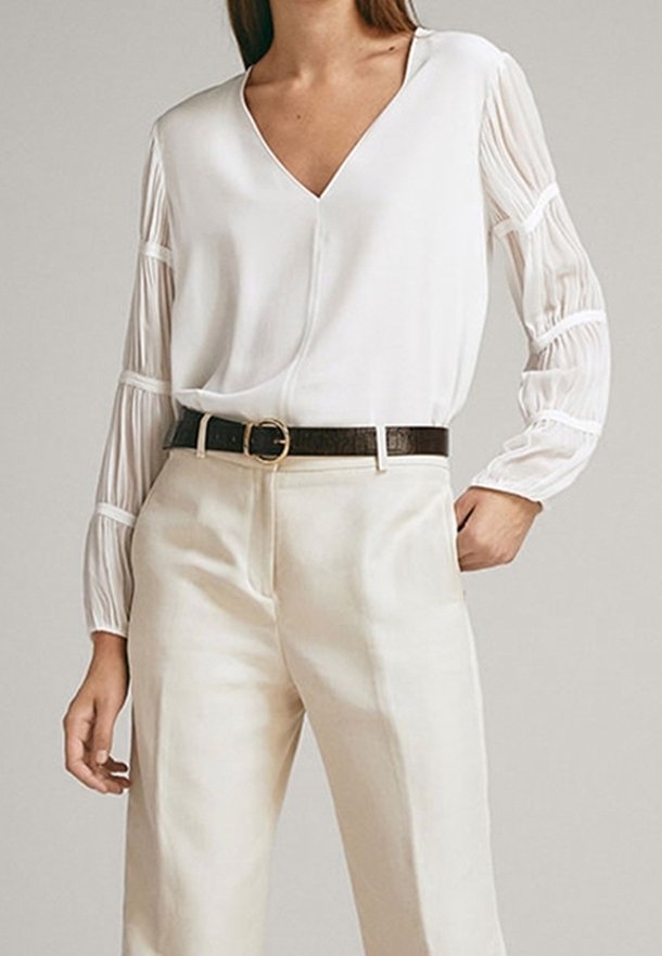 Femme portant un chemisier blanc à col en V avec des manches longues transparentes rayées et un pantalon crème taille haute avec une ceinture noire.
