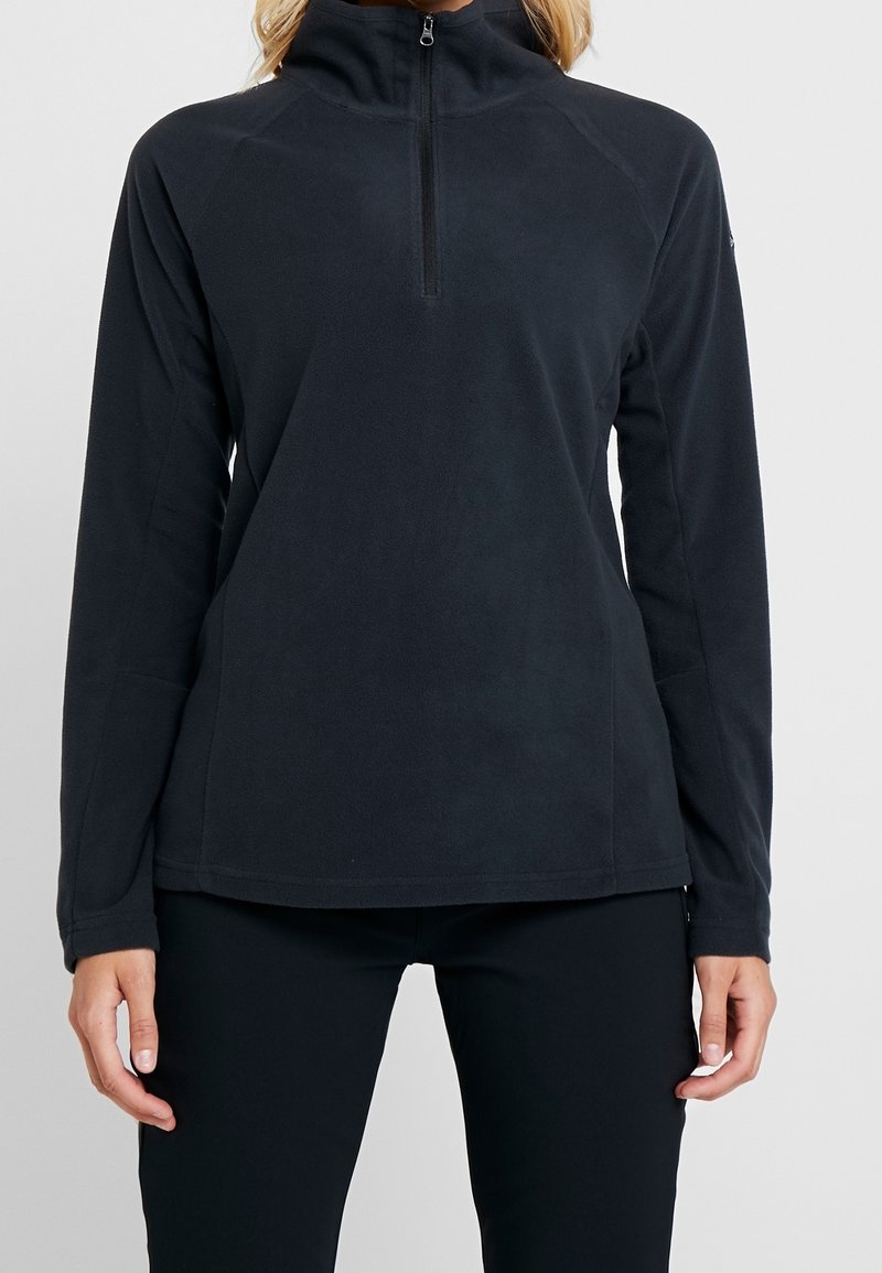 Femme portant un pull polaire noir à manches longues avec col demi-zip, assorti à un pantalon noir, debout devant un fond uni.