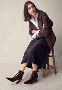 Brauner Wildledermantel über einem weißen Hemd, schwarzem Satinstück und dunkelbraunen, krokodilgemusterten Stiefeletten mit Blockabsatz.