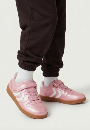 Persoon draagt roze Hummel sneakers met witte chevronstrepen, witte sokken en donkerbruine joggerbroek tegen een egale achtergrond.