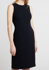 Svart ärmlös klänning med en figurnära design, slät yta, knälång fåll och subtil sömdetaljering. Inga synliga mönster eller accenter.