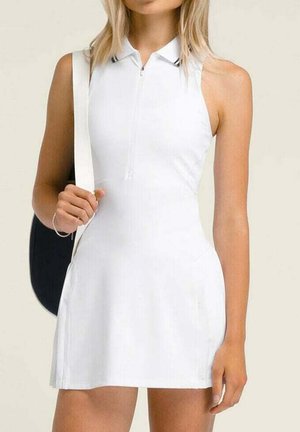 Donna che indossa un vestito da tennis bianco senza maniche con cerniera frontale, con una borsa nera a tracolla.