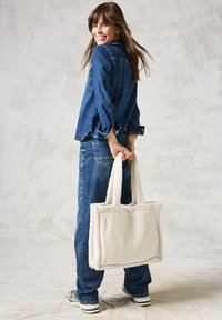 Witte, fluffy tote bag van stevig materiaal met dubbele handvatten, gecombineerd met een blauwe denimjas en jeans, casual gedragen door een model.