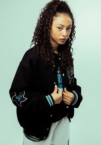 Chaqueta de béisbol negra con letras blancas y un diseño en la manga, combinada con una sudadera gráfica azul, llevada con pantalones de chándal gris claro.