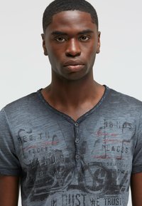 Dark gray t-shirt with a V-neck and short sleeves features graphic text in red and black. It has a slightly worn texture and two button closure.