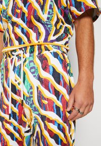 Tenue multicolore présentant un motif abstrait audacieux avec des vagues jaunes, rouges, violettes et vertes ; fabriquée en tissu léger. Taille élastique avec cordon de serrage.