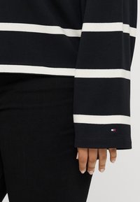 Marineblå langærmet top med hvide vandrette striber, blødt bomuldsstof og et lille mærke flag ved ærmeafslutningen.