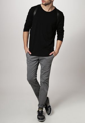 Mężczyzna ubrany w czarną koszulę z długim rękawem, szare spodnie i czarne trampki, stoi z rękami w kieszeniach i skrzyżowanymi nogami na jednolitym tle.