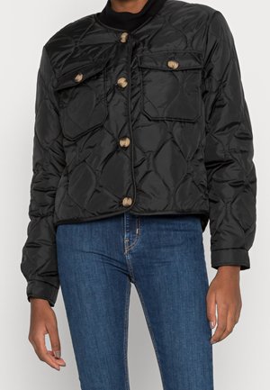 Black quilted jacket with wooden buttons and flap pockets worn with blue denim jeans on person against plain background.