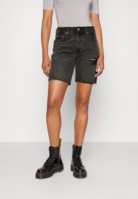 Schwarze Denim-Shorts mit ausgefranstem Saum und knielangem Schnitt. Verfügt über einen used-Look mit einem kleinen Riss am rechten Bein. Kombiniert mit schwarzen Stiefeln.