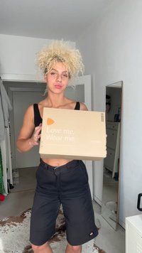 Suuri tanvärinen nelikulmainen laatikko, jossa on oranssi logo ja teksti "Rakasta minua. Pue minut päälle.", jota henkilö pitää kädessään minimalistisessa sisustuksessa, taustalla peili.