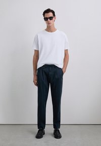 Camiseta blanca de manga corta, pantalones de pinzas azul marino y zapatos negros. El modelo está de pie contra un fondo gris claro. Gafas de sol.