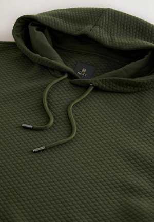 Groene gewatteerde capuchontrui met een gestructureerd oppervlak, voorzien van een trekkoord met metalen uiteinden en een label dat vlakbij de halslijn is gestikt.