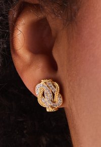 Goudkleurige oorbellen met een gedraaide vorm en heldere rhinestones, die ingewikkeld metaalwerk en gladde texturen benadrukken.