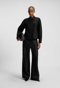 Persona che indossa una camicia nera con bottoni e pantaloni palloncino in satin, tiene una borsa nera, in piedi contro uno sfondo grigio chiaro uniforme.