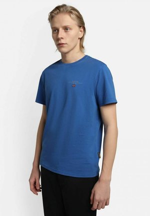 T-shirt in cotone blu con collo rotondo e maniche corte. Presenta un piccolo logo e una grafica a bandiera sul petto. Texture liscia.