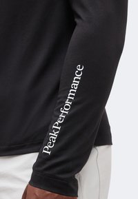 Μαύρο πουκάμισο με μακριά μανίκια με λευκό κείμενο "PeakPerformance" τυπωμένο κάθετα στον πήχη, φορεμένο από άτομο με σκούρο δέρμα και ανοιχτόχρωμο παντελόνι.