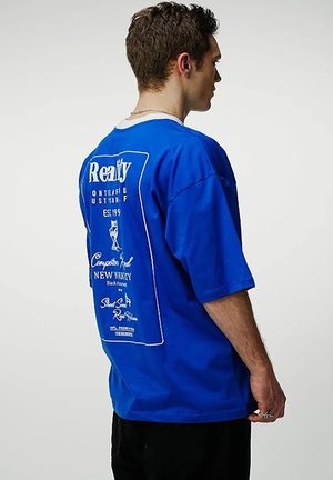 Vyras trumpais plaukais dėvintis ryškiai mėlyną labai didelio dydžio marškinėlį su baltu užrašu ir grafika ant nugaros, žiūrint iš šono.