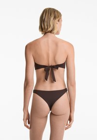 Rjav bikini komplet z bandeau zgornjim delom, ki ima vezanje na hrbtu, in nizkima hlačkami. Tkanina je gladka, v enotni barvi in minimalno oblikovana.