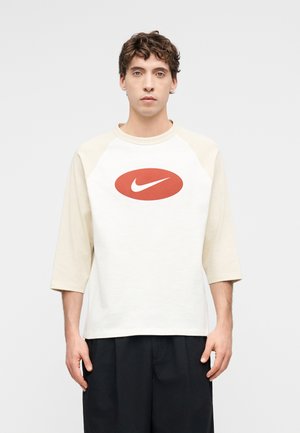 Νέος άνδρας με σγουρά μαλλιά που φοράει λευκό και μπεζ μπλουζάκι raglan μανίκια με κόκκινο οβάλ και λευκό λογότυπο Nike swoosh.