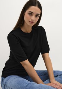 Top corto a maniche corte nero in maglia con scollatura a girocollo, texture liscia, indossato con jeans azzurri chiari, che mostrano una vestibilità rilassata.