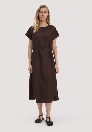 Frau in einem dunkelbraunen Midikleid mit kurzen Ärmeln, einem Taillengürtel und schwarzen geschlossenen Sandalen, stehend vor einem schlichten Hintergrund.