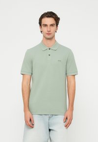 Jasnozielona koszulka polo z krótkim rękawem, kołnierzykiem i dwukolorową plisą z guzikami; z małym haftowanym logo na piersi. Noszona z jasnymi dżinsami.