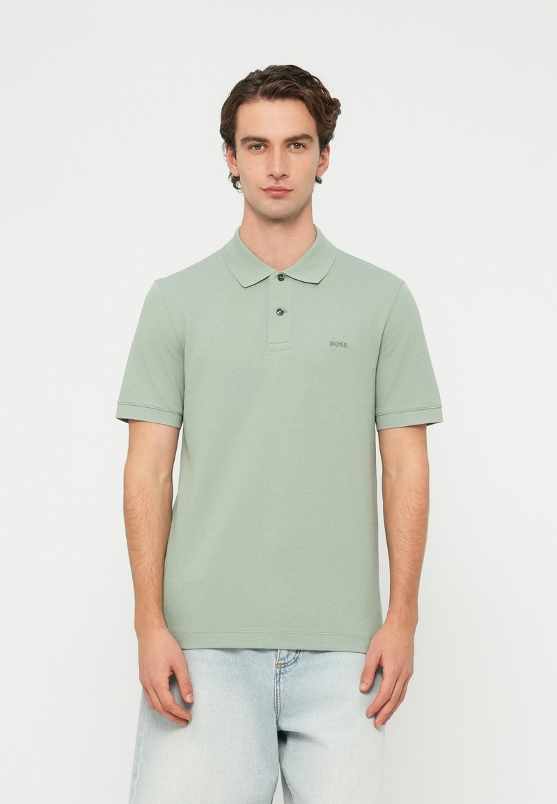 Jasnozielona koszulka polo z krótkim rękawem, kołnierzykiem i dwukolorową plisą z guzikami; z małym haftowanym logo na piersi. Noszona z jasnymi dżinsami.