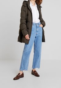 Olivengrøn pufferjakke, hvid skjorte, lyseblå jeans med høj talje og brune loafers med spænde, står på en ensfarvet baggrund.