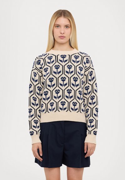 Pull en maille crème avec un motif floral hexagonal bleu marine, col rond et poignets côtelés. Associé à un short foncé.