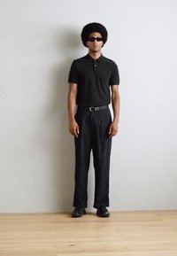 Svart polo-skjorte, svart stripete bukser og svarte sko. Bært med solbriller. Modellen har en stor, krøllete afro-frisyre. Enkel bakgrunn.