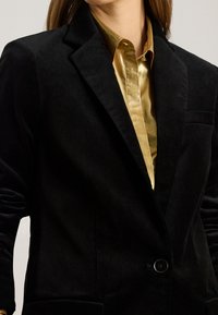 Giacca in velluto nero con lapels strutturati e chiusura a un bottone, indossata con una camicia in seta dorata lucida sotto.