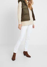 En puffer väst i olivgrön över en krämfärgad chunky knit tröja, kombinerad med vita åtsittande jeans och bruna ankelstövlar. Texturerade material är tydliga.