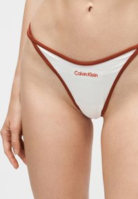 Bas de bikini blanc avec bordure couleur rouille porté sur les hanches d'une personne, avec le logo Calvin Klein au centre devant.