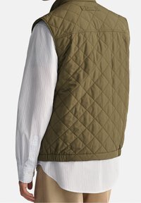 Olive green quilted vest with a high collar, side slits, and elastic hem, worn over a white striped long-sleeve shirt.