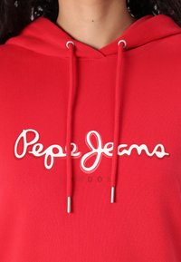 Sarkana kapuce ar auklām, ar baltu kontrastējošu logotipu, kas lasa "Pepe Jeans LONDON" augšpusē. Audums izskatās gluds un elastīgs.