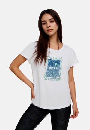 Junge Frau mit langen dunklen Haaren, die ein weißes Grafik-T-Shirt mit der Aufschrift "ONLY PLAY" und schwarze gemusterte Leggings trägt, steht mit der Hand in der Hüfte.
