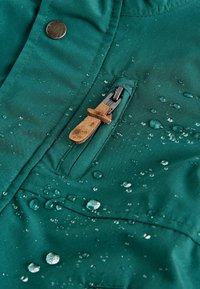 Veste imperméable verte avec une poche zippée, des boutons-pression en métal et des gouttes d'eau visibles sur la surface du tissu, mettant en valeur sa texture résistante à l'eau.